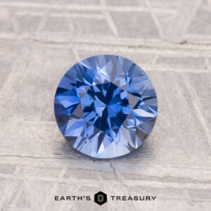A 1.48-carat heated Ceylon sapphire with a round cut is showcased on a textured grey surface, with "EARTH'S TREASURY" displayed at the bottom.