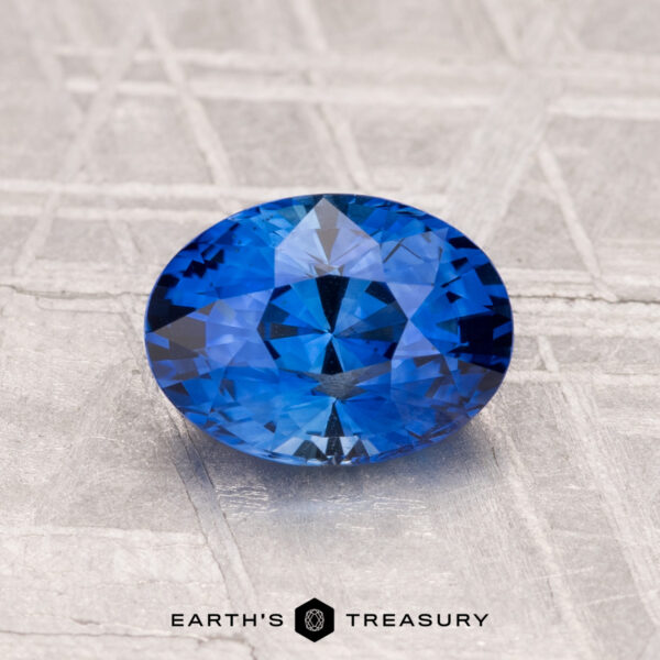 A 1.45-carat heated Ceylon sapphire, faceted in an oval shape, rests on a textured metallic surface with "EARTH'S TREASURY" displayed at the bottom.
