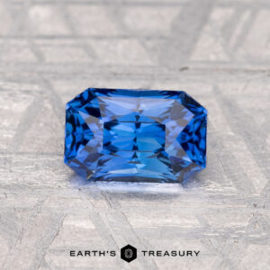 A rectangular, faceted blue 1.35-carat Ceylon sapphire (heated) rests on a textured metallic surface with "EARTH'S TREASURY" at the bottom.
