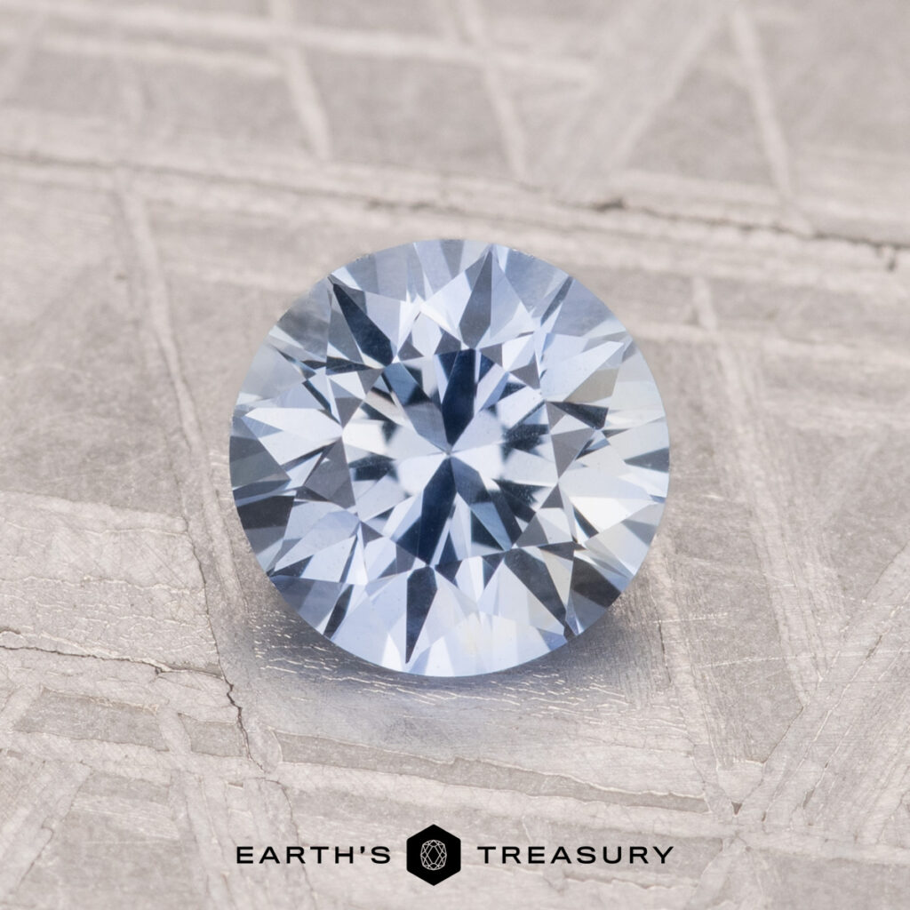A 1.28-carat heated Ceylon sapphire with a round brilliant cut and light blue hue rests on a textured gray surface beside the Earth's Treasury logo.