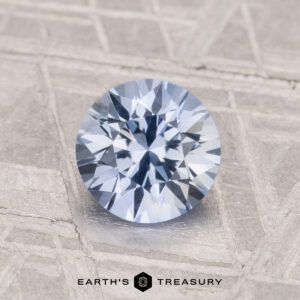 A 1.28-carat heated Ceylon sapphire with a round brilliant cut and light blue hue rests on a textured gray surface beside the Earth's Treasury logo.