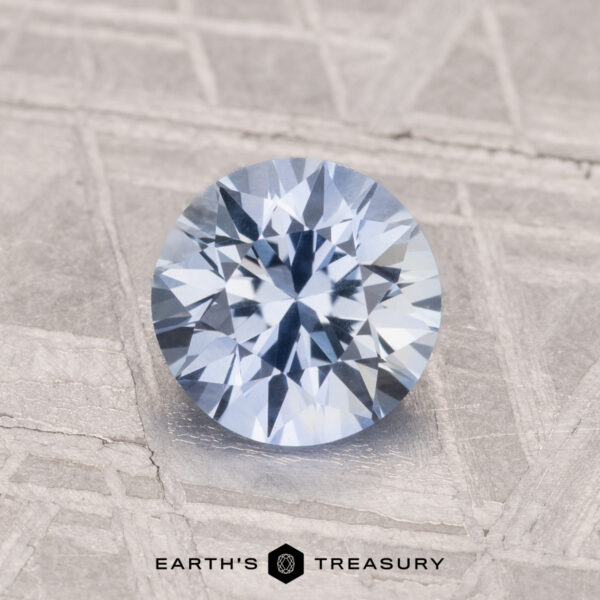 A 1.28-carat heated Ceylon sapphire with a round brilliant cut and light blue hue rests on a textured gray surface beside the Earth's Treasury logo.