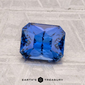 A 1.63-carat heated Ceylon sapphire, rectangular and deep blue with beveled edges, rests on a textured gray surface. "EARTH'S TREASURY" is displayed at the bottom of the image.