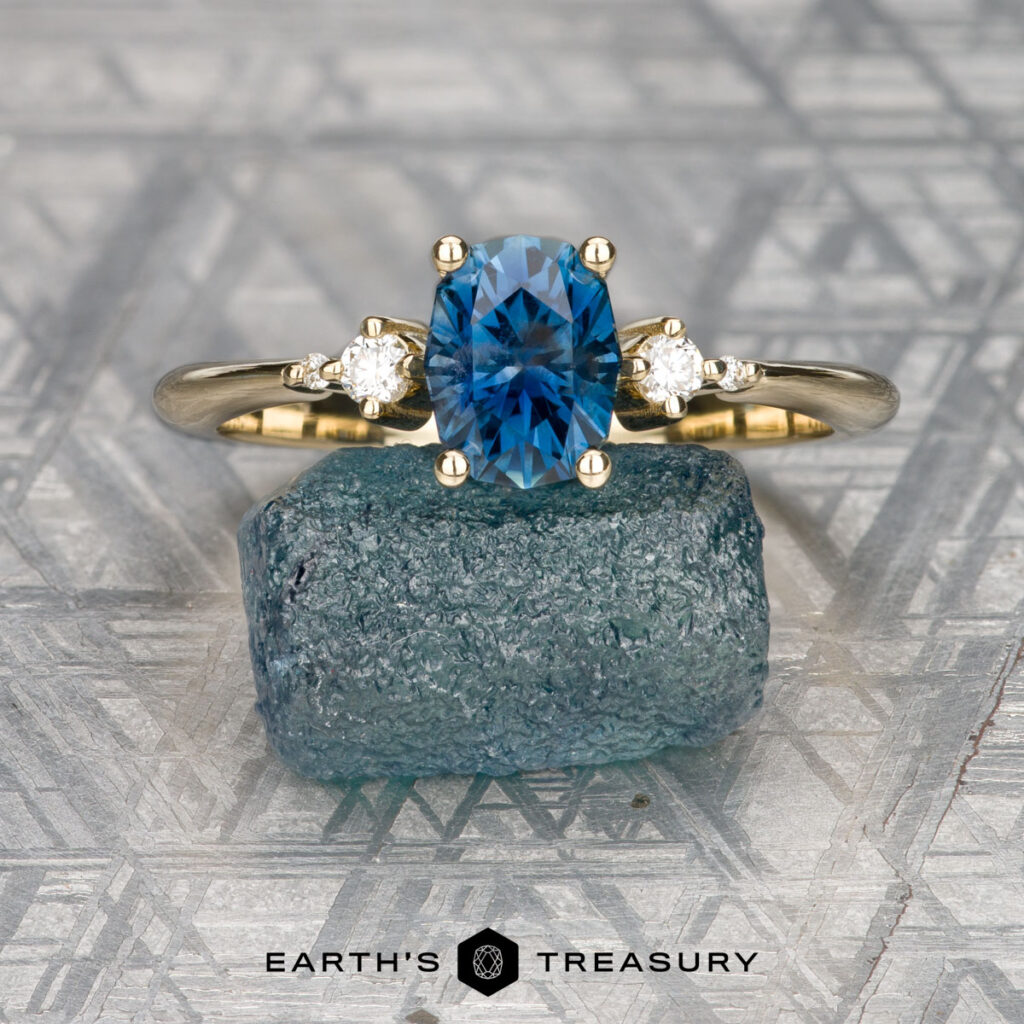 The "Selene" ring in 14k yellow gold features a 1.24-carat Montana sapphire and two small diamonds, displayed upright on a textured blue stone with a geometric patterned background. Refer to our Gem Size Guide for gemstone dimensions.