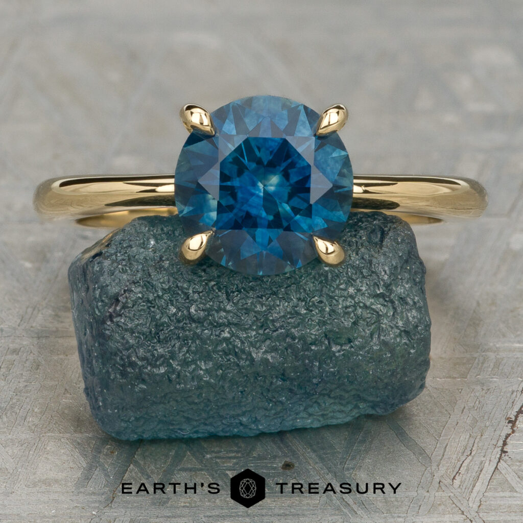 The "Sturiale, Samantha (Sam) and Rayner, Jack" gold ring with a round blue gemstone rests on a rough blue-green mineral, above the "Earth's Treasury" logo—a stylish reminder to care for your jewelry.