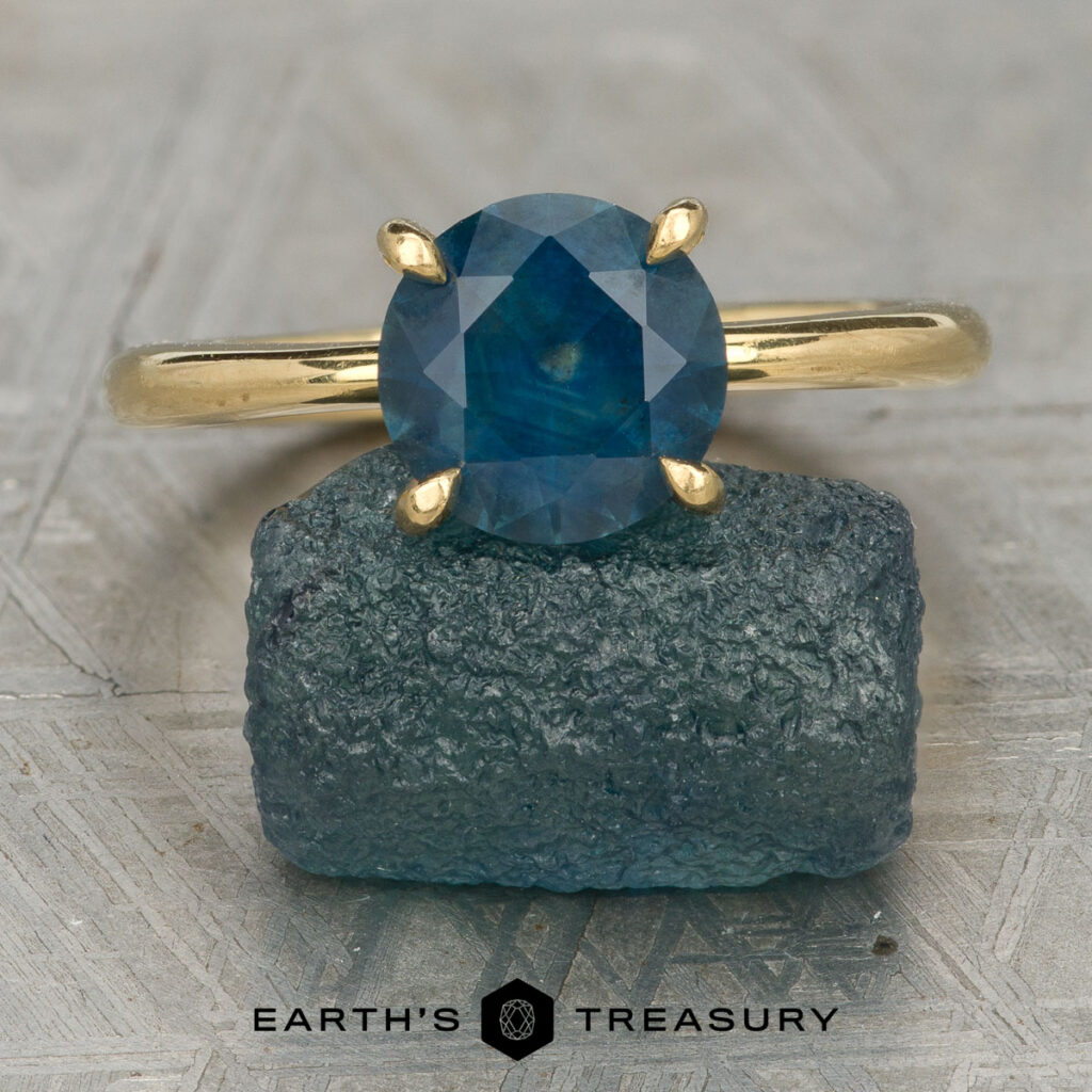 Stacked from 15 images. Method=B (R=14,S=2)" gold ring with a round blue gemstone is displayed upright on a rough blue stone. The "Earth's Treasury" logo below emphasizes caring for your jewelry to preserve its lasting brilliance.