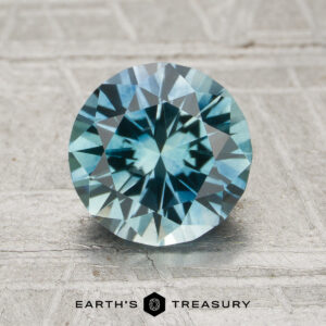 A round brilliant-cut Montana Sapphire (MS-009-250) displayed on a textured surface, featuring the "EARTH'S TREASURY" logo at the bottom.