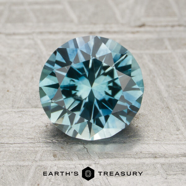 A round brilliant-cut Montana Sapphire (MS-009-250) displayed on a textured surface, featuring the "EARTH'S TREASURY" logo at the bottom.
