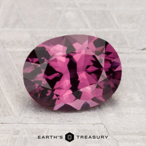 A 2.48-carat, oval-cut, deep pink spinel rests on a textured gray surface, with "Earth's Treasury" displayed below.