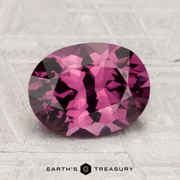 A 2.48-carat, oval-cut, deep pink spinel rests on a textured gray surface, with "Earth's Treasury" displayed below.