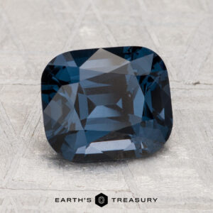 A 2.74-carat, cushion-cut deep blue spinel is displayed on a light, textured surface with the text “EARTH'S TREASURY” at the bottom.