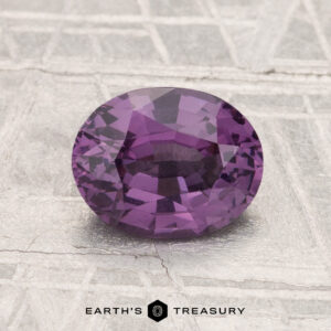 A 1.52-carat oval-cut spinel gemstone is displayed on a textured light grey surface with "EARTH'S TREASURY" below.