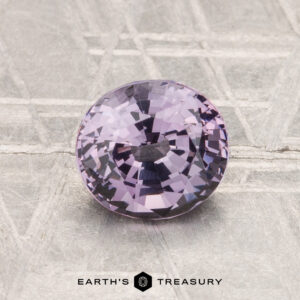A 1.35-carat, round faceted light purple spinel rests on a textured metallic surface with the text "EARTH'S TREASURY" below it.