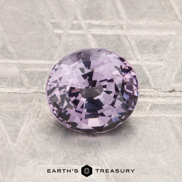 1.35-Carat Spinel A 1.35-carat, round faceted light purple spinel rests on a textured metallic surface with the text "EARTH'S TREASURY" below it.