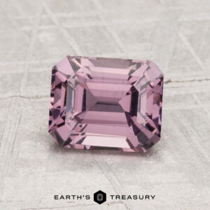 A 1.69-carat, rectangular faceted light purple spinel is displayed on a textured gray surface with the text "EARTH'S TREASURY" below it.