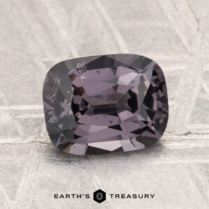 A 1.82-carat cushion-cut spinel with a purple-gray hue is displayed on a metallic, textured surface, with the text "EARTH'S TREASURY" visible at the bottom.