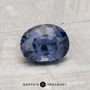 A faceted oval 1.29-carat Spinel gemstone resting on a light textured surface, with "EARTH'S TREASURY" text and logo below.