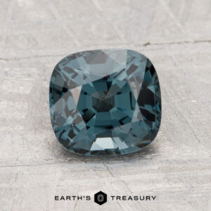 A 1.83-carat cushion-cut, deep blue-green spinel gemstone displayed on a textured grey surface with the "Earth's Treasury" logo and text below.