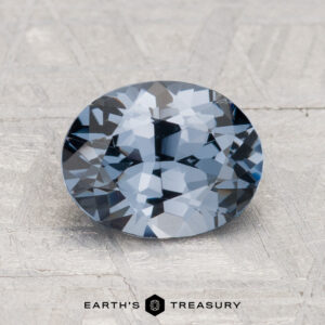 A 1.89-carat round-cut blue-gray spinel shown on a textured light gray surface with the text "Earth's Treasury" below.