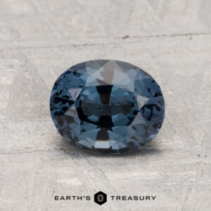 A 1.49-carat round, faceted blue spinel is displayed on a textured light gray surface with the text "EARTH'S TREASURY" below.
