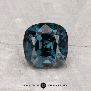 A 1.70-carat cushion-cut deep blue spinel is displayed on a textured gray surface, with the text "EARTH'S TREASURY" below.