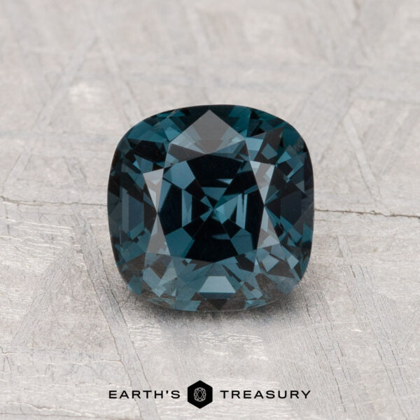 A 1.70-carat cushion-cut deep blue spinel is displayed on a textured gray surface, with the text "EARTH'S TREASURY" below.