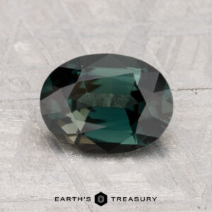 A 1.55-carat oval-cut green spinel rests on a textured light gray surface, with "EARTH'S TREASURY" displayed at the bottom.