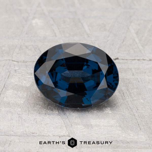 1.42-Carat Spinel A 1.42-carat oval-cut deep blue Spinel gemstone rests on a textured gray surface with "EARTH'S TREASURY" below.