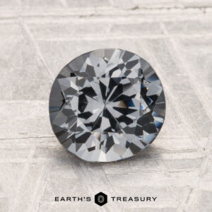 A round brilliant-cut gray 1.52-carat spinel is displayed upright on a textured surface, with the text "EARTH'S TREASURY" below it.