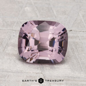 A 2.10-carat cushion-cut spinel in light purple is shown on a textured gray surface with "EARTH'S TREASURY" displayed below.
