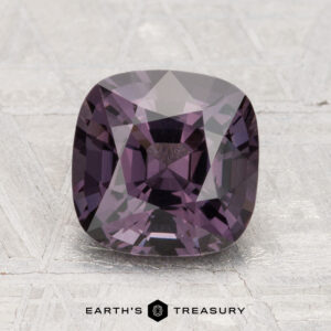 A 2.65-carat cushion-cut purple spinel is displayed on a textured gray surface, with the text "EARTH'S TREASURY" at the bottom.
