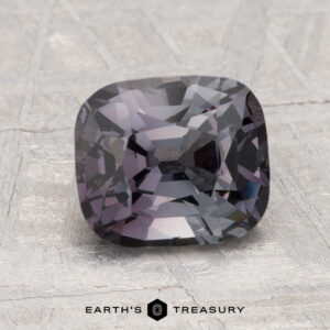 A 2.25-carat cushion-cut spinel in deep grayish-purple rests on a textured light gray surface, with "EARTH'S TREASURY" at the bottom.