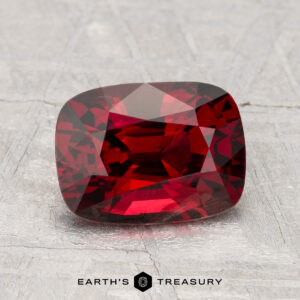 A 2.33-carat cushion-cut red Spinel is displayed on a textured light surface, with the "Earth's Treasury" logo visible at the bottom.