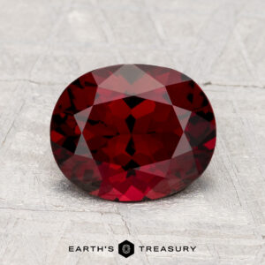 A faceted, oval-shaped 2.04-carat Spinel gemstone rests on a textured light gray surface with the text "EARTH'S TREASURY" below.