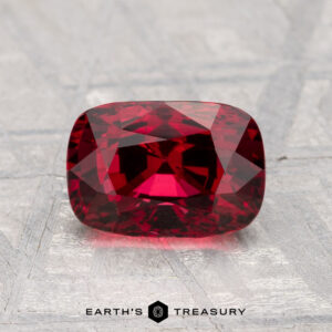 A 2.15-carat cushion-cut spinel in deep red rests on a textured silvery surface, with the "EARTH'S TREASURY" logo and text shown below.