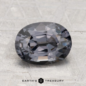 A 2.43-carat, oval-cut dark gray Spinel displayed on a textured light surface with “EARTH'S TREASURY” below.