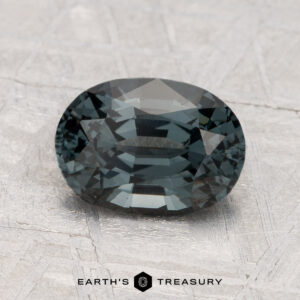 A 2.15-carat spinel gemstone, oval-cut and dark blue-green, rests on a textured light gray surface with "Earth's Treasury" written below.