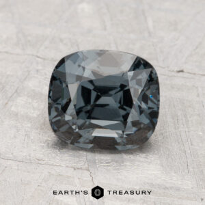 A faceted, cushion-cut 2.05-carat Spinel in dark gray is displayed on a textured gray surface with “Earth’s Treasury” written below.