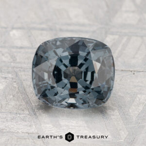A 2.19-carat, cushion-cut dark gray spinel rests on a textured surface with "EARTH'S TREASURY" displayed beneath.