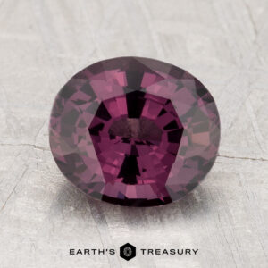 A 2.63-carat round faceted purple spinel is displayed on a light textured surface with "EARTH'S TREASURY" written below.