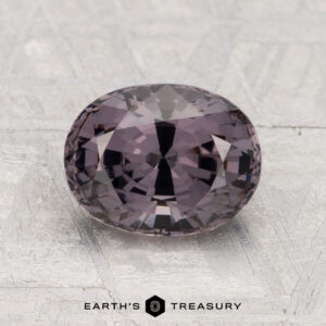 A 2.29-carat, oval-cut faceted purple spinel rests on a textured gray surface with "EARTH'S TREASURY" at the bottom.