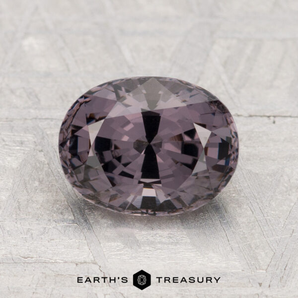 2.29-Carat Spinel A 2.29-carat, oval-cut faceted purple spinel rests on a textured gray surface with "EARTH'S TREASURY" at the bottom.