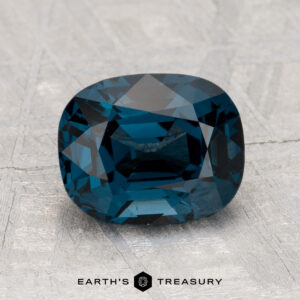 A 2.48-carat cushion-cut blue Spinel rests on a textured gray surface, with the "EARTH'S TREASURY" logo at the bottom.