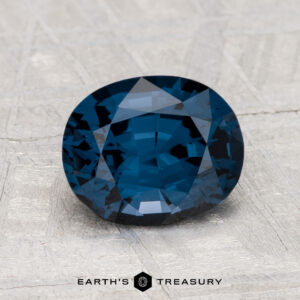 A 2.00-carat, deep blue oval-cut spinel is displayed on a textured light gray surface with the text "EARTH'S TREASURY" beneath it.