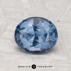 A 2.04-carat oval-cut blue spinel with multiple facets rests on a textured light gray surface. The text "EARTH'S TREASURY" appears at the bottom.