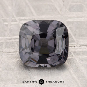 A 2.39-carat cushion-cut gray spinel displayed on a textured light gray surface, with the text "EARTH'S TREASURY" at the bottom.