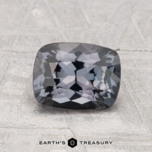 A 1.63-carat cushion-cut dark gray Spinel is shown on a textured light gray surface with "EARTH'S TREASURY" text at the bottom.