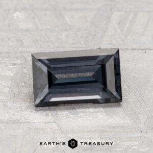 A 1.53-carat, dark blue rectangular step-cut spinel is displayed on a textured light gray surface with "EARTH'S TREASURY" text at the bottom.