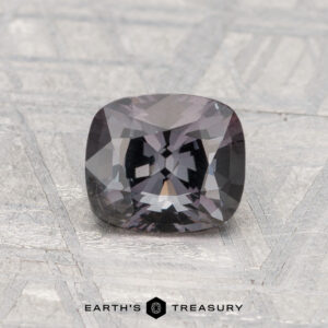 A 1.30-carat cushion-cut spinel in dark gray rests on a textured light gray surface, with "EARTH'S TREASURY" inscribed at the bottom.