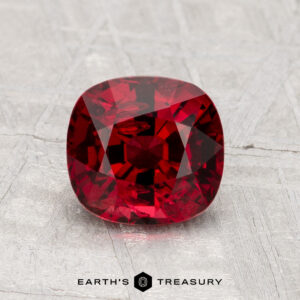 A cushion-cut 1.62-carat deep red spinel gemstone is displayed on a textured gray surface with the words "Earth's Treasury" at the bottom.